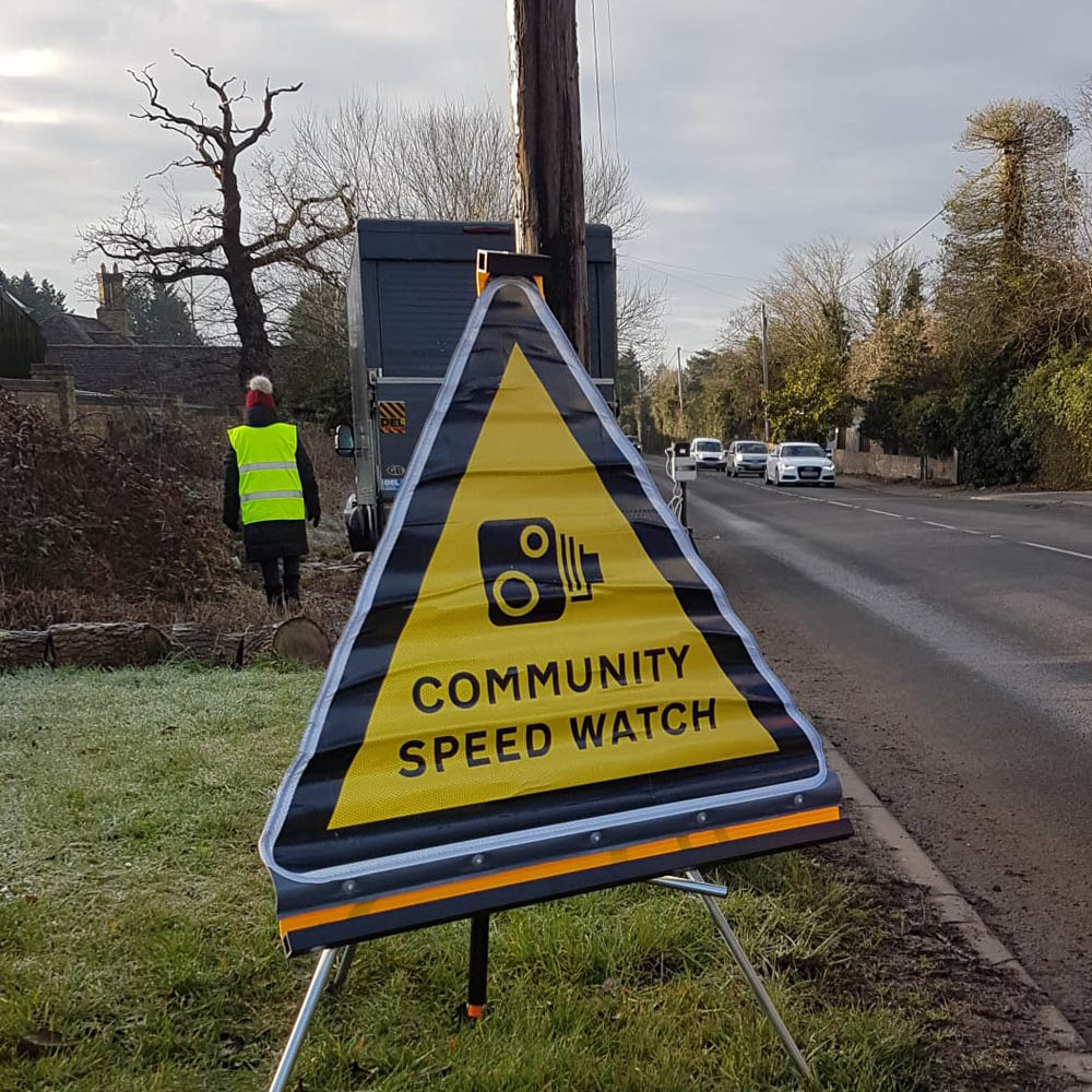 A triangular Community Speedwatch sign on a grass verge beside a road, with cars in the distance